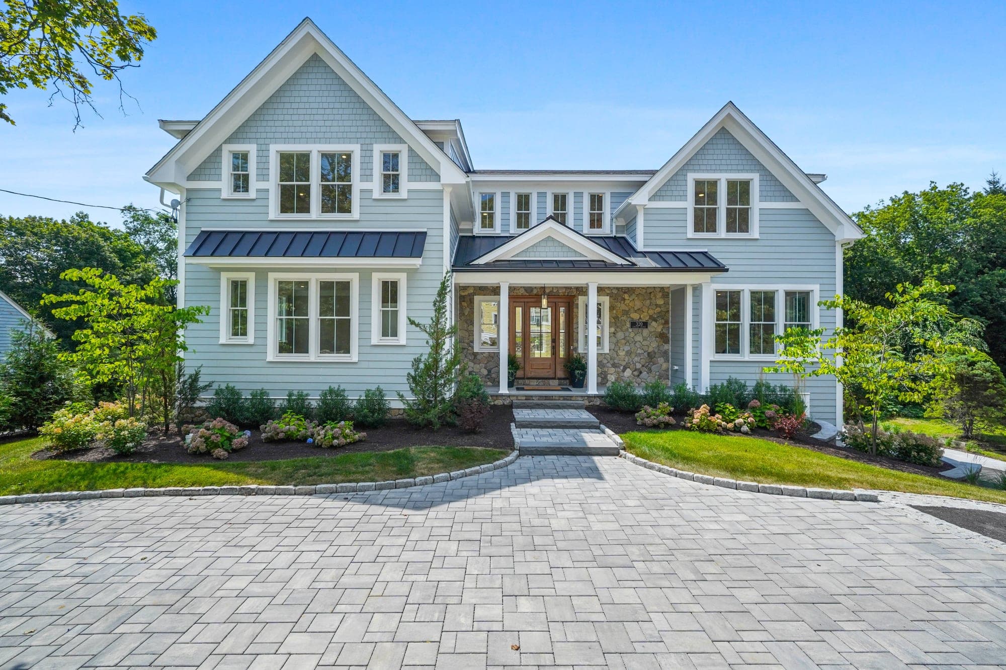 Straight-on front elevation of the light gray shingle-and-clapboard facade with stone entry, mahogany door, paver motor court, and hydrangeas in full summer bloom