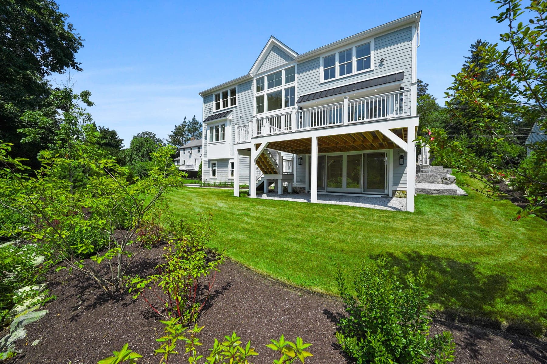 Rear three-quarter view showing the elevated deck, covered patio below, and landscaped lawn backing to woods